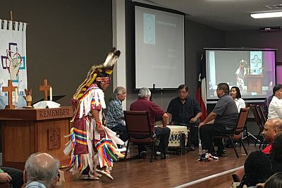 dancer in native clothing on stage, next to musicians sitting around a large drum.