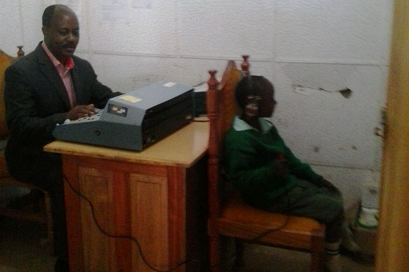 a man sits a desk, operating a hearing test machine, a child sits in front of him.